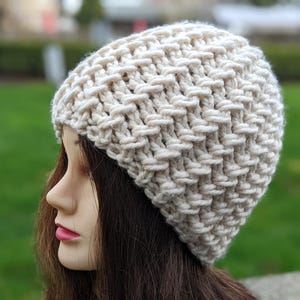 Puede incluir: Un gorro de punto de color crema con un patrón de punto texturizado. El gorro se lleva en una cabeza de maniquí con el pelo largo y castaño.