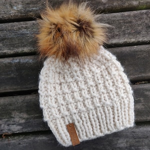 May include: A white knitted beanie with a brown faux fur pom pom. The beanie has a brown leather tag with the word "Knit" on it.