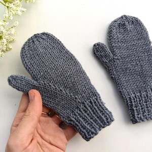 May include: A pair of gray knitted mittens. The mittens are made of a soft, textured yarn and feature a ribbed cuff. The thumb is separate from the other fingers. The mittens are displayed on a white surface.
