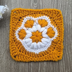 Puede incluir: Un cuadrado tejido a crochet con un diseño de flor blanca en el centro. La flor está rodeada por un borde de puntos de crochet amarillos.