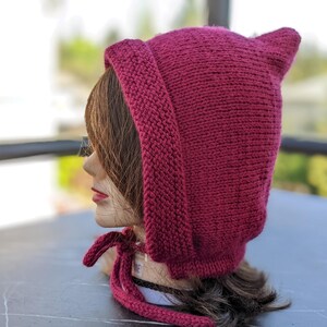 Może przedstawiać: Dzianinowy bordowy kaptur ze spiczastymi uszami i wiązanym zapięciem. Kaptur wykonany jest z grubej, teksturowanej przędzy i jest prezentowany na głowie manekina. Tło jest rozmyte, sugerując otoczenie zewnętrzne.