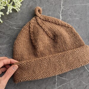 May include: A knitted beanie in a warm brown color. The hat features a folded brim and a small loop at the top. The beanie is made of a textured yarn and is shown on a gray surface with white flowers.