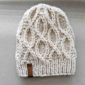 Op de afbeelding: Een crèmekleurige gebreide beanie met een kabelbreipatroon. De beanie heeft een bruin leren label met de tekst "Made with Love" erop.