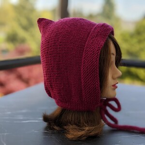 Może przedstawiać: Dzianinowy bordowy kaptur ze spiczastymi uszami, wiązany pasującym sznurkiem. Kaptur jest prezentowany na głowie manekina, prezentując teksturę i kształt ręcznie robionego akcesorium. Tło jest rozmyte, z widoczną zielenią.