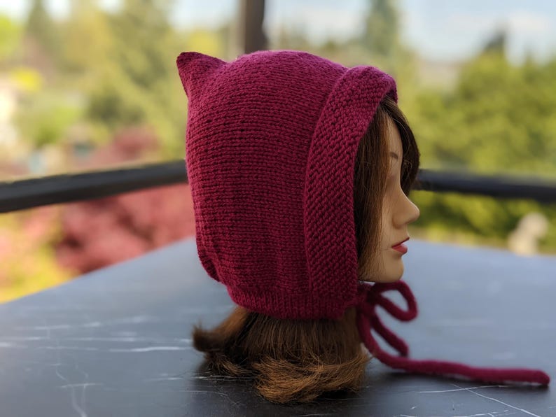 Może przedstawiać: Dzianinowy bordowy kaptur ze spiczastymi uszami, wiązany pasującym sznurkiem. Kaptur jest prezentowany na głowie manekina, prezentując teksturę i kształt ręcznie robionego akcesorium. Tło jest rozmyte, z widoczną zielenią.