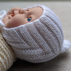 Może przedstawiać: Biały dzianinowy czapeczka dla niemowląt z małą kokardką na górze. Czapeczka jest wykonana prostym ściegiem i ma spiczasty wierzchołek.