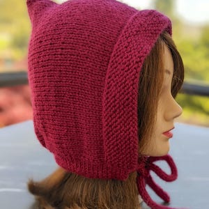 Może przedstawiać: Dzianinowy bordowy kaptur z uszopodobnymi punktami na g&oacute;rze i długimi wiązaniami. Kaptur wykonany jest z grubej, teksturowanej przędzy i ma zakrywać głowę i szyję. Wiązania służą do zabezpieczenia kaptura pod brodą.