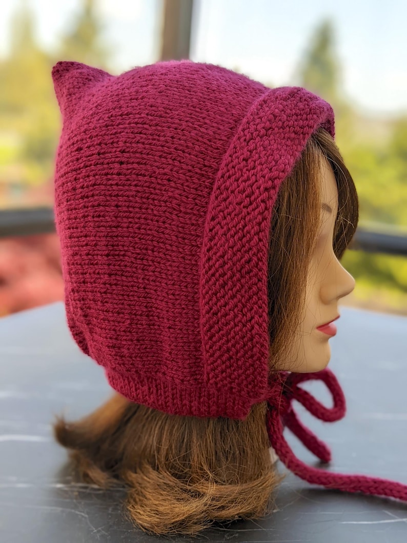 Może przedstawiać: Dzianinowy bordowy kaptur z uszopodobnymi punktami na g&oacute;rze i długimi wiązaniami. Kaptur wykonany jest z grubej, teksturowanej przędzy i ma zakrywać głowę i szyję. Wiązania służą do zabezpieczenia kaptura pod brodą.