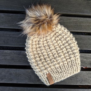 Può includere: Un berretto di lana beige con un pon pon di pelliccia sintetica marrone. Il berretto ha un'etichetta in pelle marrone con il testo "Jill" su di essa.
