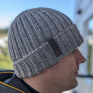 May include: A gray crocheted beanie with a brown leather patch. The beanie has a ribbed texture and a fold-over brim.