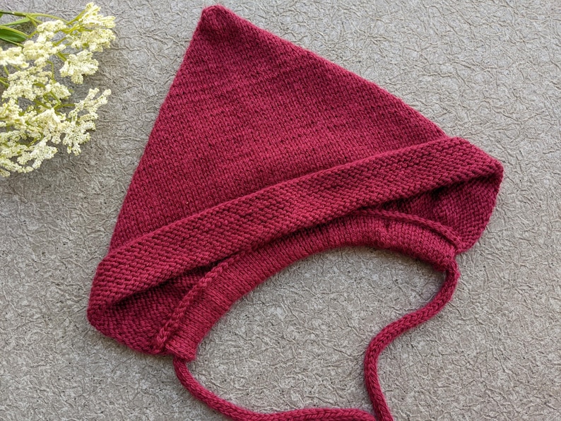 Może przedstawiać: Dzianinowa bordowa czapeczka dla dziecka z nausznikami i wiązaniami. Czapeczka wykonana jest z miękkiej, teksturowanej przędzy i ma spiczasty wierzch. Czapeczka leży na teksturowanej szarej powierzchni, z gałązką białych kwiat&oacute;w.