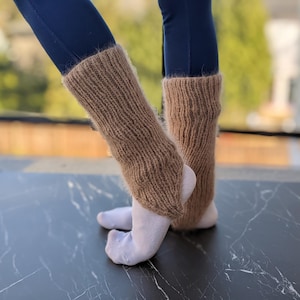 May include: A pair of brown knitted leg warmers, worn over white socks, with a ribbed texture. The leg warmers are shown against a background of blue leggings and a marble-patterned surface.