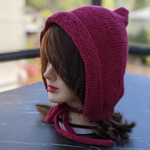 Może przedstawiać: Dzianinowa bordowa czapka z uszami i kapturem, wiązana pasującym sznurkiem. Czapka jest prezentowana na głowie manekina z brązowymi włosami i czerwoną szminką. Widoczna jest faktura czapki.