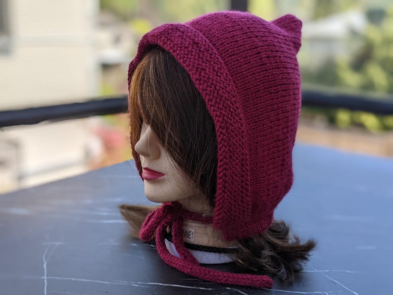 Może przedstawiać: Dzianinowa bordowa czapka z uszami i kapturem, wiązana pasującym sznurkiem. Czapka jest prezentowana na głowie manekina z brązowymi włosami i czerwoną szminką. Widoczna jest faktura czapki.