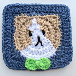 May include: A square, handmade crochet coaster featuring a dog face design. The coaster has a blue border, a tan and white face, black eyes and nose, and a green bow tie. This item is perfect for home decor.