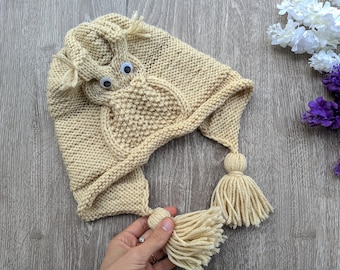 May include: A beige knitted hat with an owl design and two tassels. The owl has large white eyes.