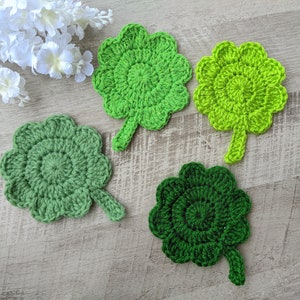 May include: Four crocheted clover leaf coasters in shades of green. The coasters are made with a textured yarn and have a raised center.