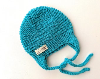 May include: A turquoise blue knitted baby hat with a tie at the back. The hat has a small tag that says "Handmade with love".