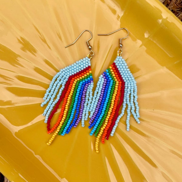 Op de afbeelding: Regenboogkleurige kralen oorbellen met een lichtblauwe achtergrond. De oorbellen zijn gemaakt van kleine kralen in een regenboogpatroon.