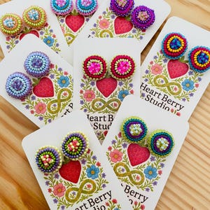 May include: A selection of beaded stud earrings in various colours, presented on white cards. Each card features a strawberry and floral design with the text "Heart Berry Studio". The earrings display combinations of pink, blue, green, and purple.
