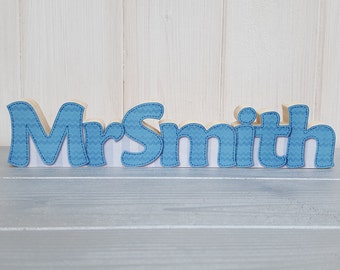 May include: Wooden sign spelling "Mr Smith" in a playful font. The letters are light wood with blue chevron patterned accents and stitched edges. The sign is displayed on a light blue surface against a white background.