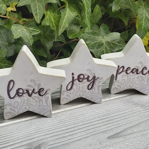 May include: Three white wooden star decorations with the words "love", "joy", and "peace" in dark brown script. Each star has a grey floral design and a distressed finish. The stars are on a grey wooden surface.