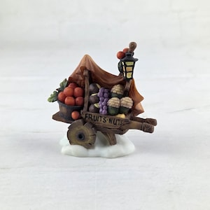 May include: A miniature fruit and nut cart figurine. The cart is made of wood and features a variety of fruits, including red apples and purple grapes. The cart has a brown canopy and a lantern. The text "FRUITS.NUTS" is visible on the cart.