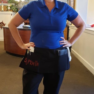 May include: A woman wearing a blue polo shirt and black pants with a black apron that says "Big Mouth" on it. She is standing in a restaurant.