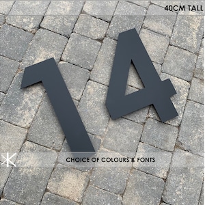 Op de afbeelding: Zwarte metalen huisnummers 1 en 4, elk 40 cm hoog, getoond op een grijs bakstenen oppervlak. De nummers zijn in een modern lettertype. De tekst "CHOICE OF COLOURS & FONTS" is zichtbaar onder de nummers.
