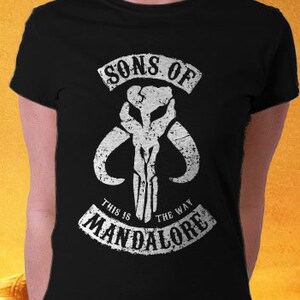 May include: Black t-shirt with a white graphic of a Mandalorian symbol. The text "Sons of Mandalore" and "This is the way" are printed on the shirt.