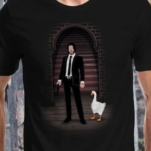 Puede incluir: Una camiseta negra con un gráfico de un hombre con traje y un ganso. El hombre sostiene una pistola y el ganso está de pie junto a él. El gráfico está sobre un fondo oscuro con una puerta y escaleras.