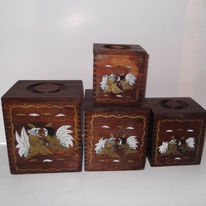 May include: A collection of four wooden boxes in a dark brown hue. Each box is adorned with a design of two birds, incorporating white and gold details. The boxes are square, with a circular aperture on the top of the two larger ones.