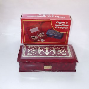 May include: A burgundy jewelry box with gold floral detailing and a matching red box with the text "Coffret à maquillage et à bijoux". The box has a rectangular shape and a small gold plaque on the front.