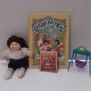 May include: A Cabbage Patch Kids doll with brown hair, wearing a white shirt, black pants, and white shoes. Beside the doll is a book titled "Cabbage Patch Kids The Great Rescue" and a miniature swing set.