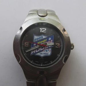 May include: A silver-toned wristwatch with a dark gray face. The watch face features the text "2005 Wrangler Nationals Rodeo Las Vegas" in blue, white, and red. The watch has a silver band and a round face with the numbers 12, 3, and 9.