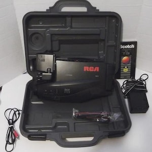 May include: A black RCA camcorder with 24X zoom, inside a grey carrying case. The case is open, revealing the camcorder, a power adapter, and a VHS tape. The RCA logo is in red. A black and red cable is also visible.
