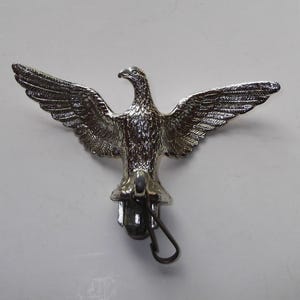 May include: Silver-colored metal eagle with outstretched wings. The eagle has detailed feathering and a hooked beak. A small loop is attached below the eagle's body. The eagle is likely a decorative item or emblem.