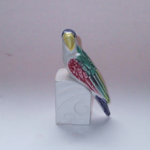 Puede incluir: Figura de loro de cerámica posada sobre un bloque blanco. El ave presenta cabeza azul, pico amarillo, alas verdes y rojas y cola azul. El bloque tiene un diseño decorativo de remolino.