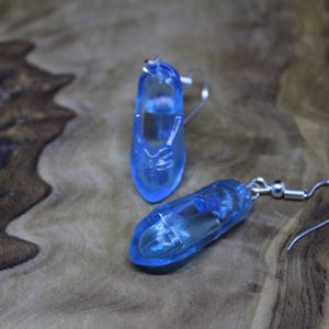 May include: A pair of translucent light blue shoe-shaped earrings. Each earring features a clear blue resin charm molded into the shape of a small shoe, attached to a silver-toned metal fishhook ear wire for pierced ears.