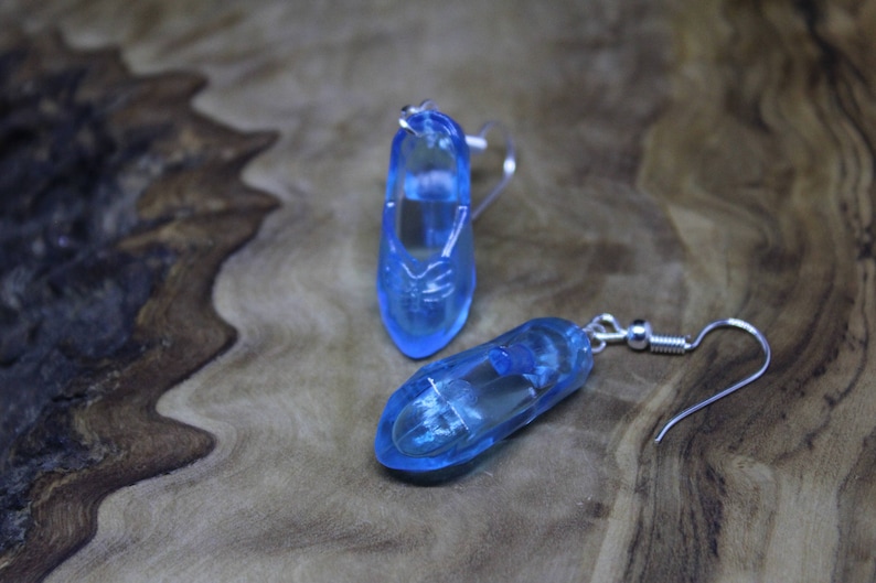 May include: A pair of translucent light blue shoe-shaped earrings. Each earring features a clear blue resin charm molded into the shape of a small shoe, attached to a silver-toned metal fishhook ear wire for pierced ears.