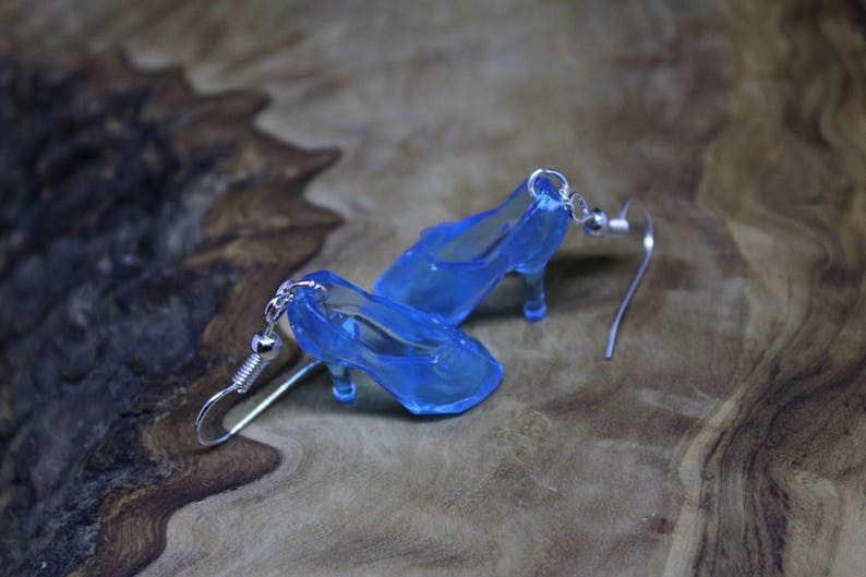 May include: A pair of dangle earrings featuring miniature translucent blue high-heeled shoe charms. Each charm is attached to a silver-toned metal fishhook ear wire. The earrings are crafted from a clear, blue-tinted material that mimics the appearance of glass, showcasing a pointed toe and a slender heel design.
