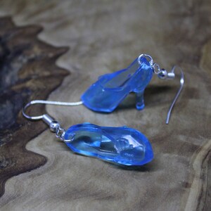 May include: A pair of dangle earrings featuring miniature translucent blue high-heeled shoe charms. Each charm is attached to a silver-toned metal fishhook ear wire with a small jump ring, creating a delicate and whimsical accessory design.