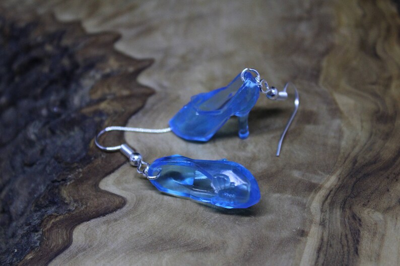 May include: A pair of dangle earrings featuring miniature translucent blue high-heeled shoe charms. Each charm is attached to a silver-toned metal fishhook ear wire with a small jump ring, creating a delicate and whimsical accessory design.