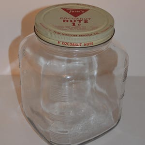 May include: A clear glass jar with a metal lid. The lid is off-white with red and black text that reads "Tom's COCOANUT HUTS 1¢". The jar has a square shape with rounded corners and vertical measurement markings. The jar is empty.