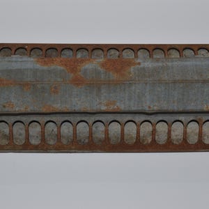 May include: A rectangular, weathered metal object with a series of oval openings along the top and bottom edges. The metal has a gray hue with patches of rust, suggesting age and exposure to the elements. The object appears to be a vintage feeder.