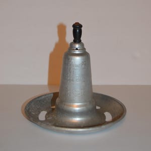 May include: A silver-colored metal poultry roaster stand with a conical center post and a wide, circular base featuring four cut-out slots. The center post has a black knob at the top and embossed markings indicating measurements for liquid capacity.
