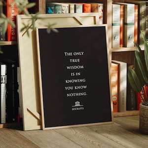 Könnte beinhalten: Ein rechteckiger schwarzer Wandkunstdruck in einem dünnen Holzrahmen mit dem weißen Text: The only true wisdom is in knowing you know nothing. Unter dem Text befindet sich ein kleines weißes Symbol eines griechischen Tempels und der Name Socrates.