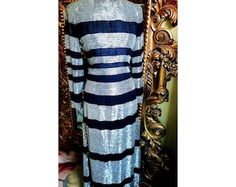 May include: A vintage silver sequin dress with navy blue horizontal stripes. The dress has long sleeves and a high neckline. The dress is displayed on a mannequin. The background includes a gold ornate mirror.