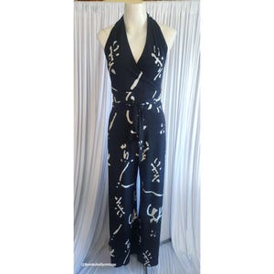 Könnte beinhalten: Schwarzer Neckholder-Jumpsuit mit weißem, abstraktem Muster. Der Jumpsuit hat ein Wickeloberteil, eine Bindetaille und eine Hose mit weitem Bein. Das Kleidungsstück wird auf einer Schaufensterpuppe vor einem weißen Vorhang präsentiert.