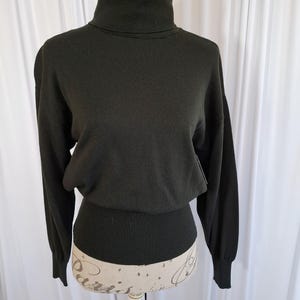 May include: A dark olive green long-sleeve sweater featuring a high turtleneck collar, dropped shoulders, and a wide ribbed knit waistband that creates a blouson effect. The garment is crafted from a smooth, solid-colored knit fabric.